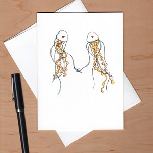 Peut inclure: Une carte blanche avec une illustration dessin&eacute;e &agrave; la main de deux m&eacute;duses avec des c&oelig;urs rouges sur la t&ecirc;te. Les m&eacute;duses se tiennent la main et ont de longues tentacules flottantes.