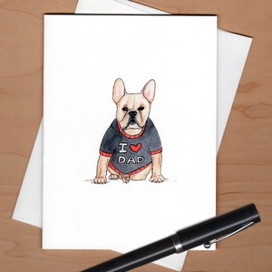 Pu&ograve; includere: Biglietto di auguri ad acquerello con un bulldog francese che indossa un maglione nero con "I ❤️ DAD" in rosso e bianco. Il biglietto &egrave; su una busta bianca, con una penna nera in primo piano. Lo sfondo &egrave; una superficie di legno chiaro.
