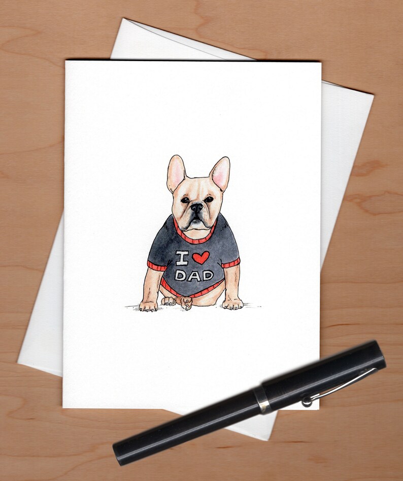 Pu&ograve; includere: Biglietto di auguri ad acquerello con un bulldog francese che indossa un maglione nero con "I ❤️ DAD" in rosso e bianco. Il biglietto &egrave; su una busta bianca, con una penna nera in primo piano. Lo sfondo &egrave; una superficie di legno chiaro.