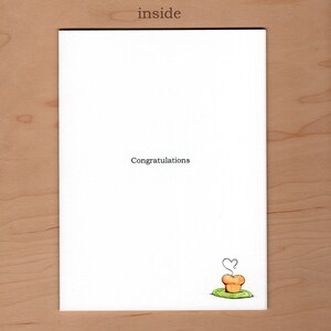 May include: A white card with the word "inside" printed at the top. The card has the text "Congratulations" printed in black ink. A small cartoon drawing of a cupcake with a heart-shaped steam rising from it is printed in the bottom right corner.