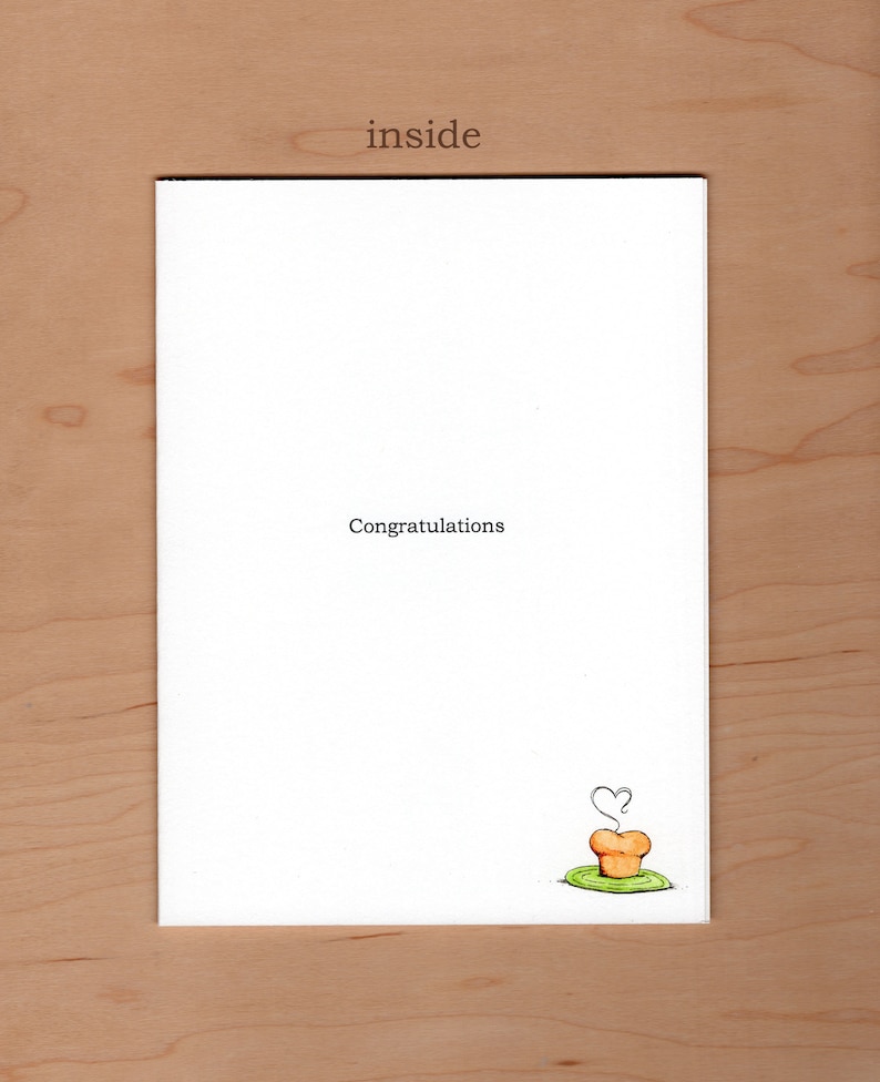 May include: A white card with the word "inside" printed at the top. The card has the text "Congratulations" printed in black ink. A small cartoon drawing of a cupcake with a heart-shaped steam rising from it is printed in the bottom right corner.