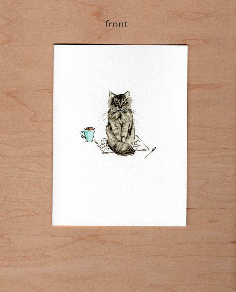 Front of greeting card;  pen and watercolor illustration of a long haired cat sitting in the middle of an open book a pen and cup sit to the side
