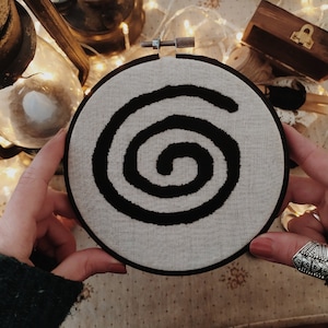 May include: A black and white embroidery hoop with a spiral design. The hoop is held in a person's hands.