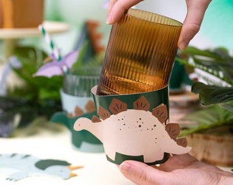 May include: A brown glass cup with a dinosaur-shaped paper cup holder. The dinosaur is green, brown, and pink with a white body. The cup holder is designed to hold a glass cup.