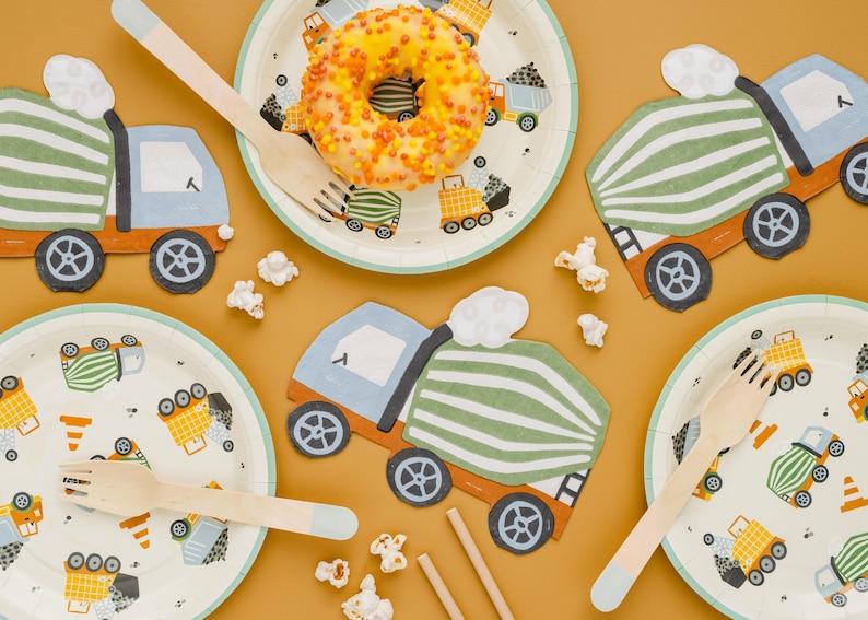 May include: A festive party scene with construction-themed decorations. Includes paper plates and truck-shaped cutouts, wooden forks, and a glazed donut. The plates feature colorful construction vehicles. Popcorn is scattered around the scene.