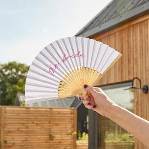 May include: White paper fan with a gold-colored wooden handle, featuring the pink text "the bride." The fan is open, displaying its pleated design. The fan is held up against a background of a wooden building and a sunny sky.