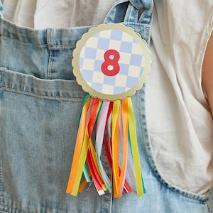 Op de afbeelding: Een ronde badge met een geschulpte gouden rand en een lichtblauw en wit geblokt patroon. Het nummer 8 is rood. Meerkleurige linten in rood, oranje, geel, groen en paars hangen eronder. De badge is vastgemaakt aan een spijkerbroek.