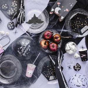 May include: A Halloween party table setting with black and white decorations, including a skeleton hand, a black cat, and a witch's hat. There are also plates, cups, and balloons with a Halloween theme.