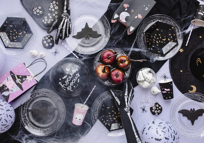 May include: A Halloween party table setting with black and white decorations, including a skeleton hand, a black cat, and a witch's hat. There are also plates, cups, and balloons with a Halloween theme.