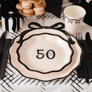 Può includere: Un'apparecchiatura per la tavola da festa a tema bianco e nero. Il centrotavola è composto da piatti con il numero "50" e la parola "hello" stampata. Ci sono anche macarons, una scatola regalo e un bicchiere di carta.