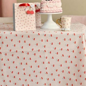 Peut inclure: Une nappe rose pâle avec un motif répétitif de cerises rouges, un sac cadeau assorti avec une anse en ruban rouge, un présentoir à gâteau blanc portant un gâteau rose givré et une tasse blanche avec un motif de cerises et une anse dorée.