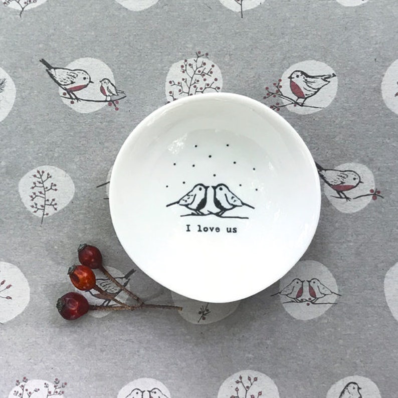 Op de afbeelding: Een wit keramiek sieradenschoteltje met een ontwerp van twee vogels die elkaar aankijken in een besneeuwde sc&egrave;ne. Het schoteltje heeft de tekst "I love us" gedrukt op de bodem.