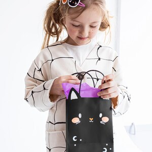 May include: A young girl wearing a white cardigan with black spiderweb design holds a black Halloween treat bag with a cat face and purple tissue paper inside. The bag has white stars and a crescent moon on it.