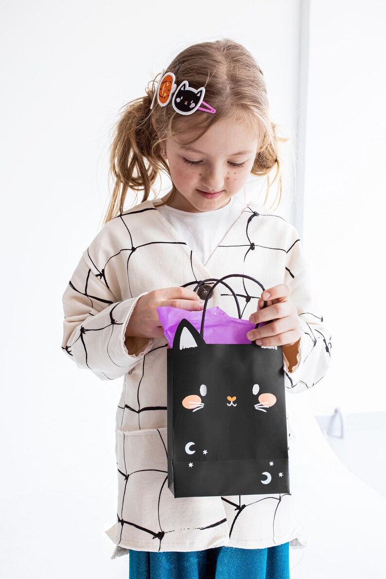 May include: A young girl wearing a white cardigan with black spiderweb design holds a black Halloween treat bag with a cat face and purple tissue paper inside. The bag has white stars and a crescent moon on it.