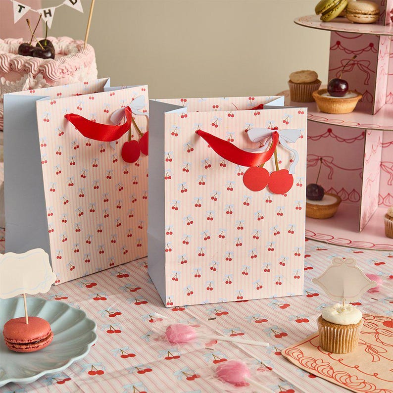 May include: Two rectangular paper gift bags with a light pink and white vertical striped pattern featuring small red cherry illustrations. Each bag has a bright red ribbon handle and a decorative light blue paper bow accent attached to the top.