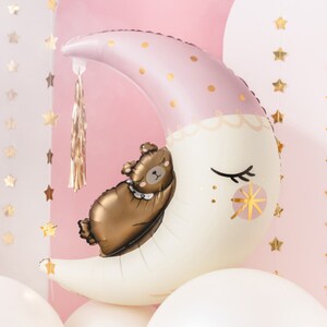 Può includere: Un palloncino a forma di luna crescente rosa e bianco con accenti di glitter dorati. Un palloncino a forma di orsacchiotto marrone dorme sulla luna. Il palloncino ha un fiocco dorato appeso.