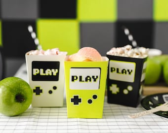 May include: Three yellow and black party favor boxes with the word "PLAY" on them. The boxes are filled with snacks and are sitting on a white and green checkered tablecloth.