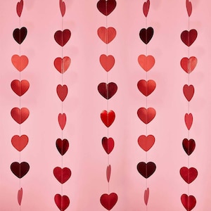 Könnte beinhalten: Ein rosa Hintergrund mit mehreren Strängen herzförmiger Dekorationen in verschiedenen Rottönen. Die Herzen sind vertikal aufgehängt und erzeugen eine festliche und romantische Atmosphäre. Ideal für Valentinstag oder andere Feiern.