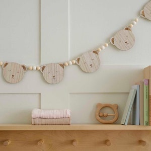 May include: A wooden bear-shaped garland with light brown beads and bear faces with closed eyes. A wooden bear-shaped rattle and a stack of pink and tan blankets are on a wooden shelf. Books are on the right.