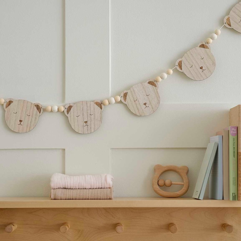 May include: A wooden bear-shaped garland with light brown beads and bear faces with closed eyes. A wooden bear-shaped rattle and a stack of pink and tan blankets are on a wooden shelf. Books are on the right.