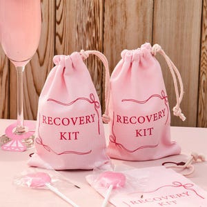 Peut inclure: Deux sacs roses à cordon avec les mots "RECOVERY KIT" en rouge. Les sacs sont sur une surface rose avec deux sucettes roses et un verre à vin rose. L'arrière-plan est un mur en bois.