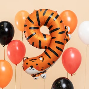 Könnte beinhalten: Orangefarbener und schwarz gestreifter Ballon in Form der Zahl neun mit einem Tigergesicht am Boden. Der Ballon ist von orangefarbenen, roten und schwarzen Ballons umgeben.