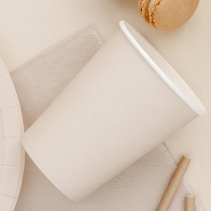 Könnte beinhalten: Ein becher aus beigem Papier mit weißem Rand liegt auf einer beigen Serviette. Ein Pappteller und ein Strohhalm sind ebenfalls sichtbar. Ein hellbraunes Makronen-Gebäck befindet sich im Hintergrund. Die Artikel sind auf einer hellbeigen Oberfläche angeordnet.