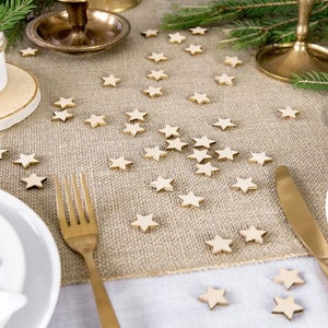 May include: A burlap table runner with scattered wooden star-shaped decorations. The table runner is set for a festive dinner with gold cutlery, white plates, and a gold candle holder.