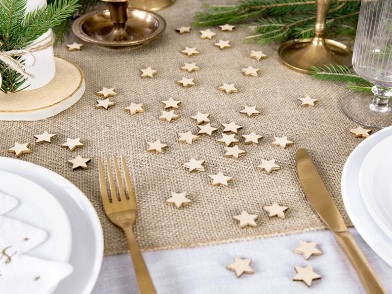 May include: A burlap table runner with scattered wooden star-shaped decorations. The table runner is set for a festive dinner with gold cutlery, white plates, and a gold candle holder.