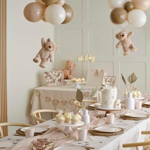 May include: A baby shower setup with a table set for a celebration. The table is decorated with a cake, cupcakes, and cookies. Teddy bear decorations and balloons in neutral colors add to the festive atmosphere. The words "Oh Baby" are in a neon sign.