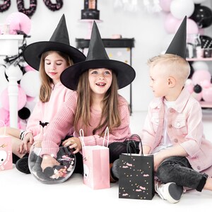May include: Three children dressed in pink and black Halloween costumes. Two girls wear black witch hats and pink sweaters. One girl holds a glass bowl filled with candy. The boy wears a black witch hat and a pink jacket. All three children hold pink and black Halloween treat bags.