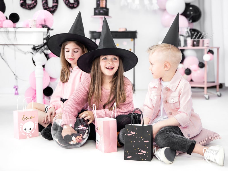 May include: Three children dressed in pink and black Halloween costumes. Two girls wear black witch hats and pink sweaters. One girl holds a glass bowl filled with candy. The boy wears a black witch hat and a pink jacket. All three children hold pink and black Halloween treat bags.