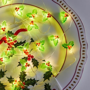 May include: A festive string of fairy lights with green holly leaves and red berries. The lights are displayed on a white plate with a red and brown patterned rim, and a holly-printed napkin.