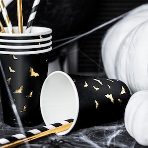 May include: Black paper cups with gold bat silhouettes, perfect for Halloween. The cups are stacked and next to a fallen cup with a black and white striped straw. White pumpkins and spiderwebs complete the spooky scene.