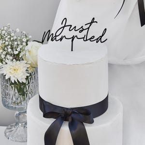 Op de afbeelding: Een witte bruidstaart met twee lagen, een zwart lint en een strik. Een zwarte caketopper leest "Just Married" in cursief schrift. Een vaas met witte bloemen is zichtbaar op de achtergrond.