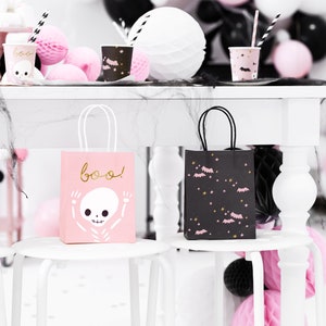 May include: Two Halloween party favor bags, one pink with a skeleton and the text "boo!" and the other black with pink bats and stars. The bags are on white stools with a white table in the background.
