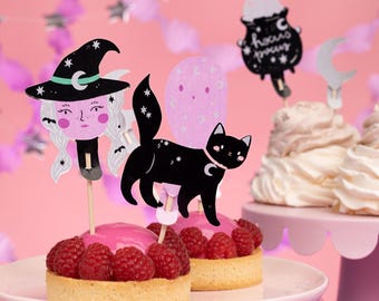 Może przedstawiać: Dwie mini tarty w kolorze różowym i białym, ozdobione malinami i dekoracjami na torty w stylu Halloween. Dekoracje to czarownica, czarny kot i duch. Tarty stoją na różowym stojaku na torty.