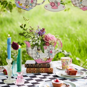May include: An Alice in Wonderland themed tea party setting. A floral arrangement in a teapot-shaped container sits on books. Teacups, plates, and candles are arranged on a black and white checkered tablecloth. Decorative teapots hang above.