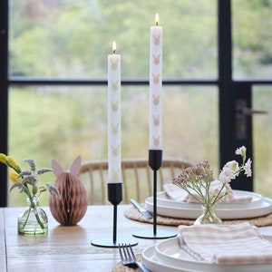 Könnte beinhalten: Zwei brennende weiße Kerzen mit einem sich wiederholenden Hasenmuster, in schwarzen Metallhaltern. Die Kerzen stehen auf einem Holztisch, zusammen mit Osterdekorationen und Blumengestecken. Die Szene spielt sich vor einem Fenster ab.