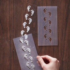 May include: A clear plastic stencil with a row of footprint shapes. A hand is holding a second clear plastic stencil with a row of footprint shapes. The stencils are on a brown wooden surface. The footprints are filled with white powder.