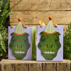 Könnte beinhalten: Zwei lavendelfarbene Geschenktüten mit grünen Dinosauriergesichtern und Partyhüten. Die Dinosauriergesichter haben weiße Zähne und gelbe Augen. Die Partyhüte sind grün und rosa gestreift mit einem orangefarbenen Pompon. Orangefarbene Schleifenhenkel.