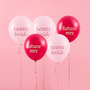Könnte beinhalten: Fünf Ballons, einige weiß und einige rot, werden vor einem rosa Hintergrund gezeigt. Die weißen Ballons tragen den Text "team bride" und die roten Ballons "future mrs". Bänder hängen von den Ballons.