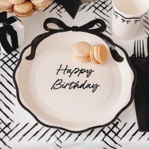 May include: A white paper plate with a black bow design and the words "Happy Birthday" in black script. Two macarons sit on the plate. The plate is part of a party setting with other black and white decorations.