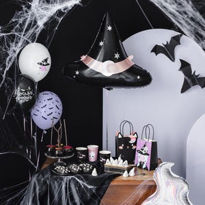 May include: A Halloween party table setting with black and purple balloons, a black witch hat balloon, and black bats. There are also black and white gift bags with Halloween characters, black and white cups, and black and white candy on a black tablecloth.