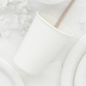 May include: A white paper cup with a wooden stir stick inside, positioned on a white surface. White paper plates and floral accents are also visible, suggesting a party or event setting.