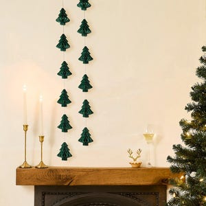 May include: A festive scene with dark green paper Christmas tree decorations hanging against a cream wall. Two lit candles in gold holders sit on a wooden mantel, with a glass of wine and a reindeer figurine.
