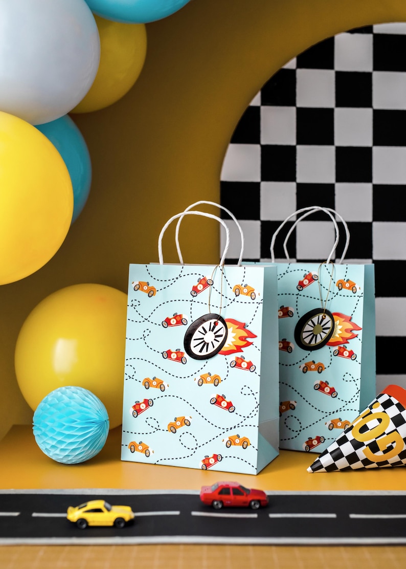 K&ouml;nnte beinhalten: Zwei hellblaue Geschenkt&uuml;ten mit wei&szlig;en Griffen sind mit orangefarbenen und roten Rennwagen und gepunkteten Linien verziert. Jede Tasche hat einen schwarzen Radanh&auml;nger mit einer roten und orangefarbenen Flamme. Gelbe und blaue Luftballons sind im Hintergrund.
