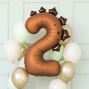 Op de afbeelding: Een bruine folieballon met het cijfer 2 en een dinosaurusrugontwerp, omringd door een cluster van ballonnen in wit, goud en lichtgroen. Dit feestelijke ballonnenarrangement is perfect voor een verjaardagsfeest.