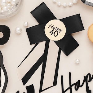 May include: A black ribbon bow with a circular sticker that reads "Happy 40" in black script. The bow is on a white background with black and white striped accents. Scattered pearls and a container of pearls are also visible.