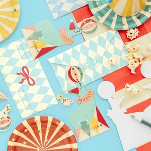 Op de afbeelding: Een set circus-thema feestbenodigdheden, waaronder borden, uitnodigingen en decoraties. De borden zijn rood en geel met een stervormig ontwerp. De uitnodigingen zijn blauw en wit met een ruitpatroon. De decoraties omvatten een blauwe en groene olifant met een rode en witte ballon, een witte olifant-uitstans en een witte kaart met het woord "hallo" erop.